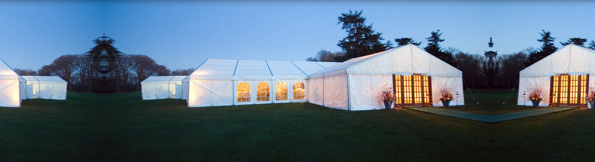 A row of lit Shipseys marquees at dusk on a lawn, framed by mature trees
